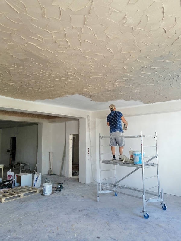Pracownik na rusztowaniu nakłada strukturalną masę na sufit w jasnym pomieszczeniu, tworząc unikalny wzór z wyraźnymi śladami pędzla.