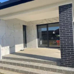 Nowoczesny taras z marmurowymi płytkami na ścianie, czarną cegłą na filarze i trzema betonowymi stopniami wejściowymi.