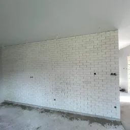 Świeżo położona, biała cegła dekoracyjna na ścianie działowej w surowym wnętrzu, widoczne otwory na gniazdka i wystające kable, surowy beton na podłodze.