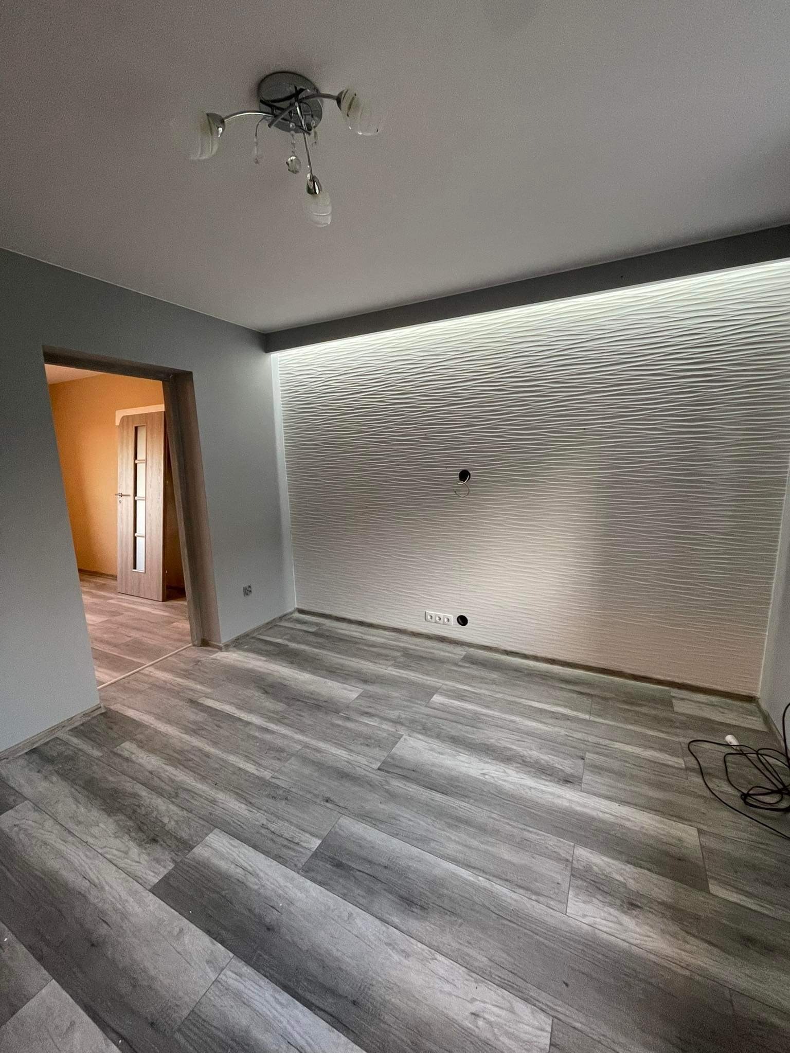 Pokój po remoncie: szare ściany, panel podłogowy imitujący drewno, dekoracyjna biała ściana z podświetleniem LED i otwory na gniazdka, widok na otwarte drzwi do innego pomieszczenia.