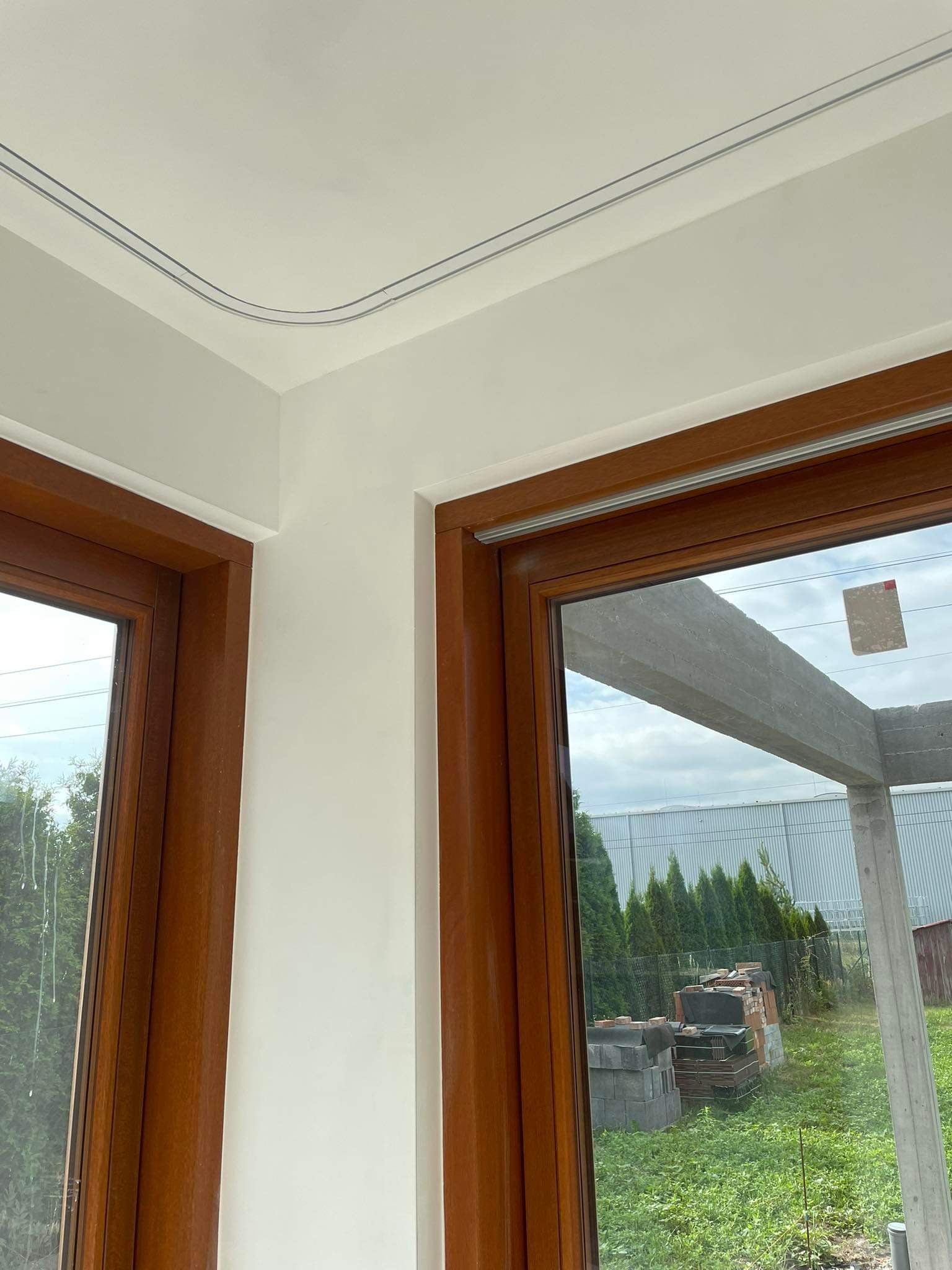 Wnętrze z dwoma drewnianymi oknami, widok na zewnątrz na teren budowy z cegłami i zielenią, pomalowane ściany na biało, dekoracyjny pasek na suficie.