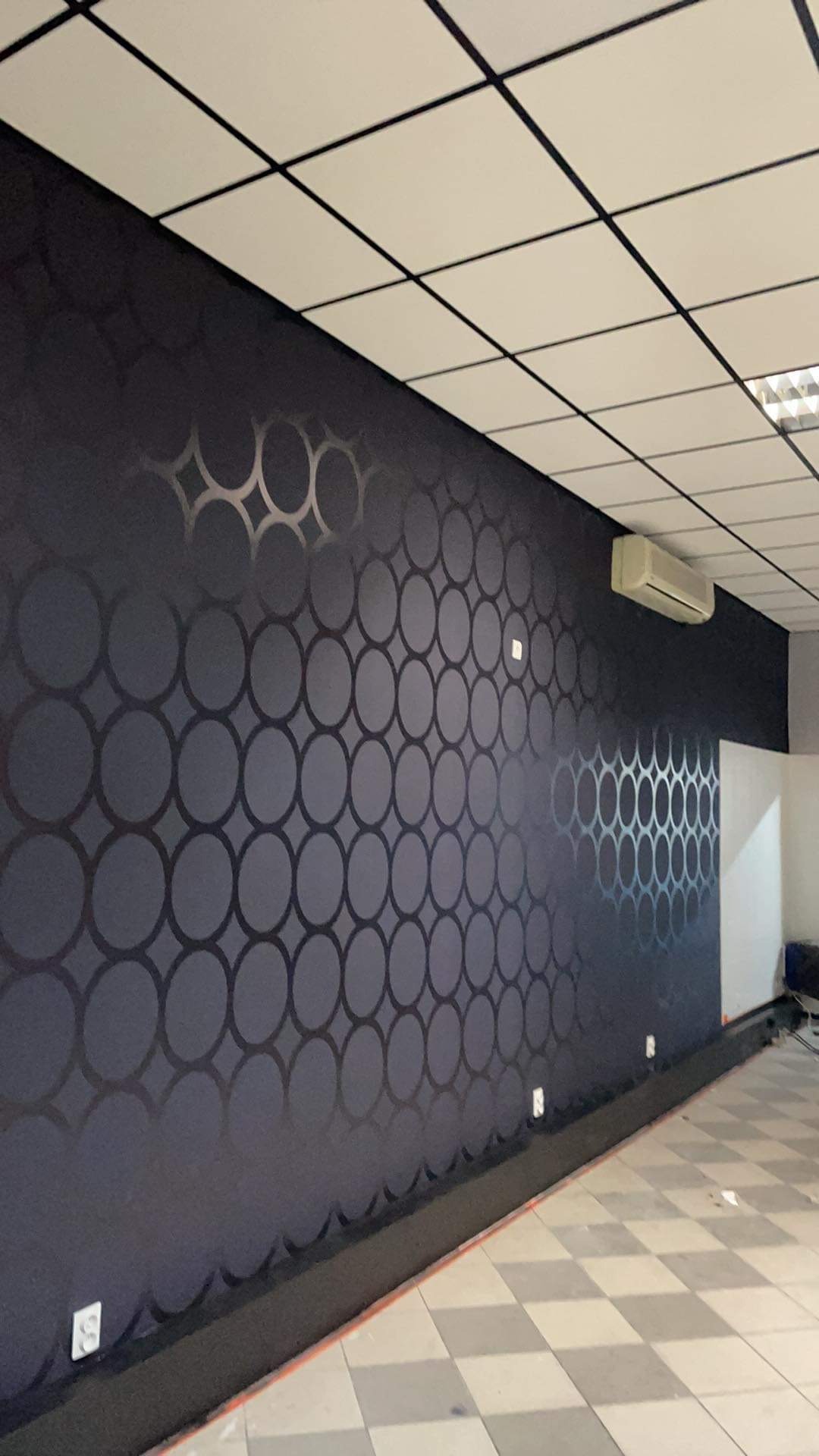 Ściana pokryta ciemną tapetą w geometryczny wzór kół, z widocznym klimatyzatorem i fragmentem szachownicowej podłogi.