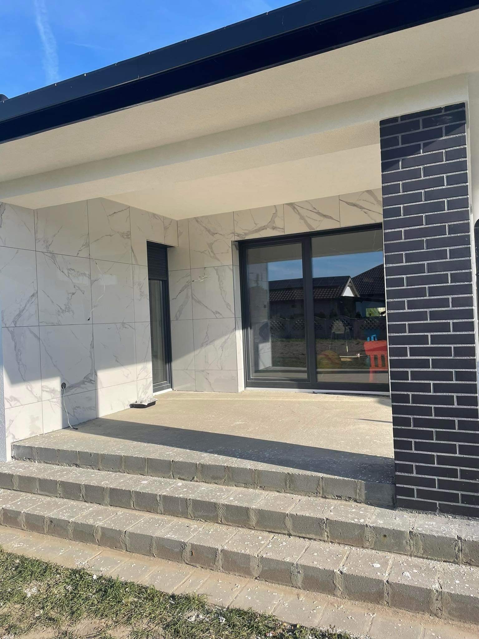 Nowoczesny taras z marmurowymi płytkami na ścianie, czarną cegłą na filarze i trzema betonowymi stopniami wejściowymi.