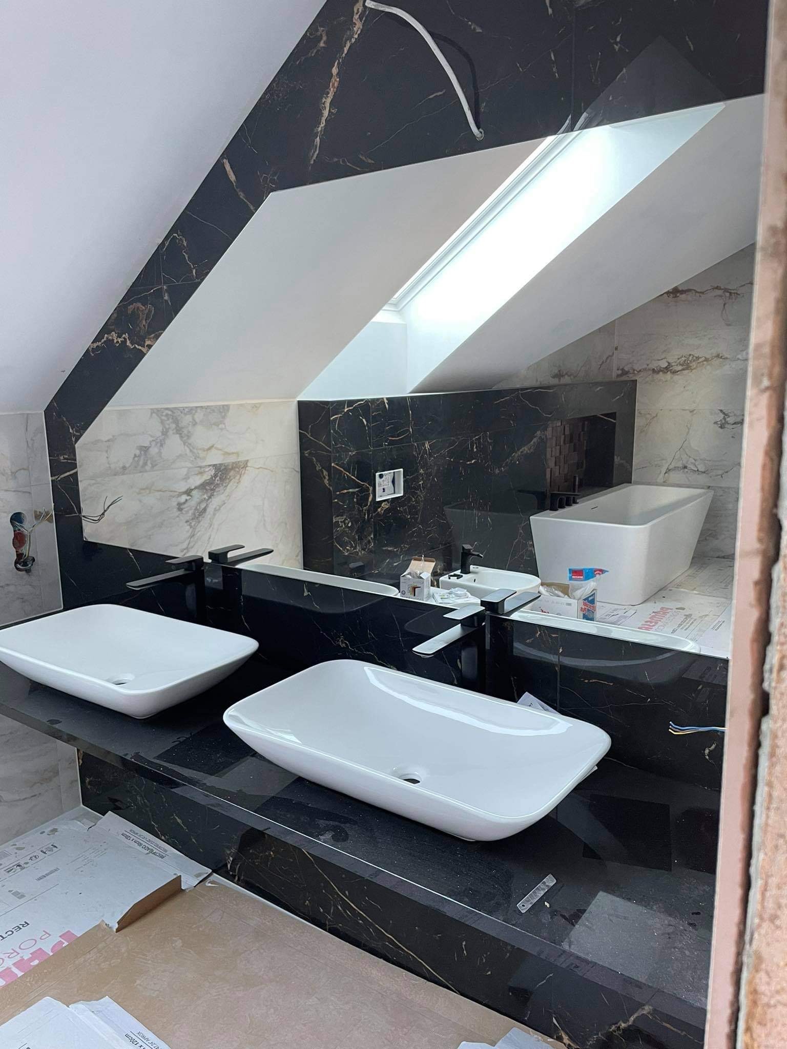 Łazienka w trakcie wykańczania z dwiema białymi umywalkami na czarnym blacie z marmurowym wzorem, widoczna wanna, okno dachowe i fragmenty płytek oraz materiałów budowlanych.