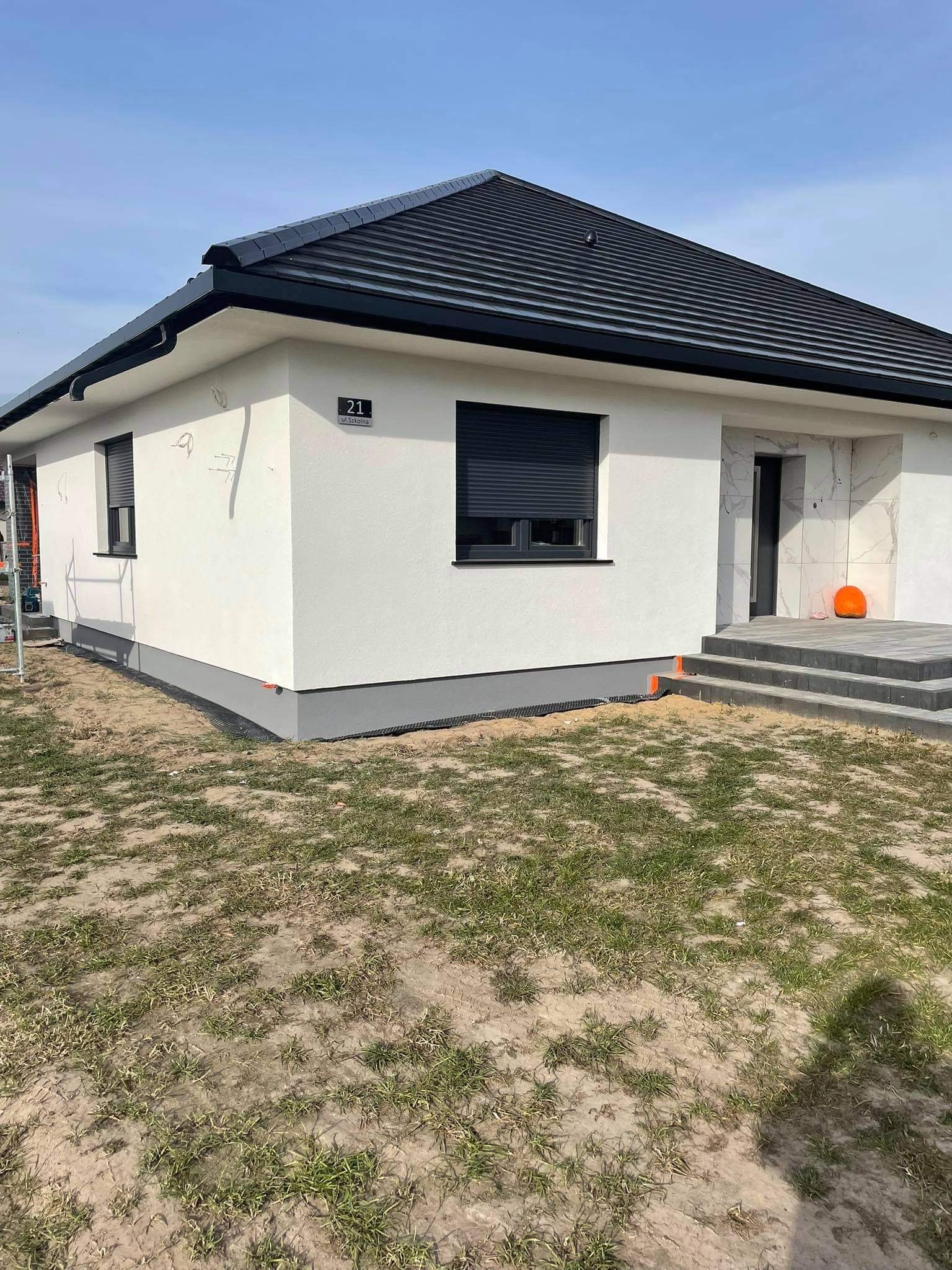 Biały dom jednorodzinny z ciemną dachówką i szarym cokołem, widoczne ślady prac wykończeniowych na elewacji, częściowo zagospodarowany teren wokół budynku.