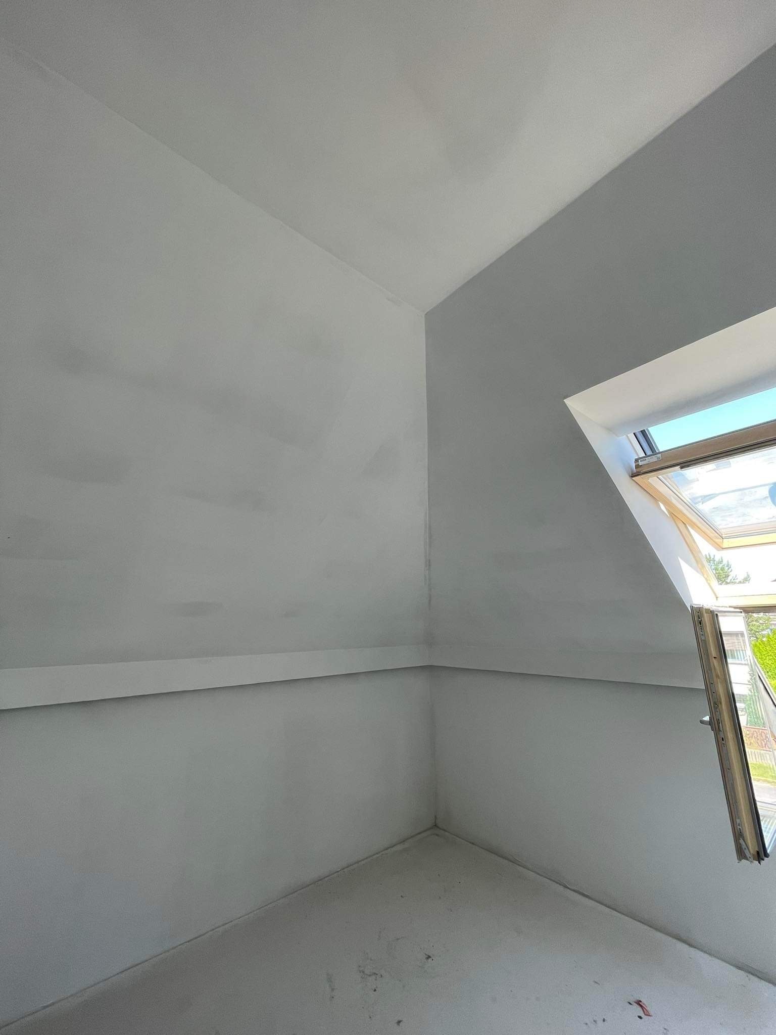 Pomalowane na szaro poddasze z oknem dachowym, widoczne niedoskonałości malowania na ścianach i suficie, ślady po taśmie malarskiej na oknie.