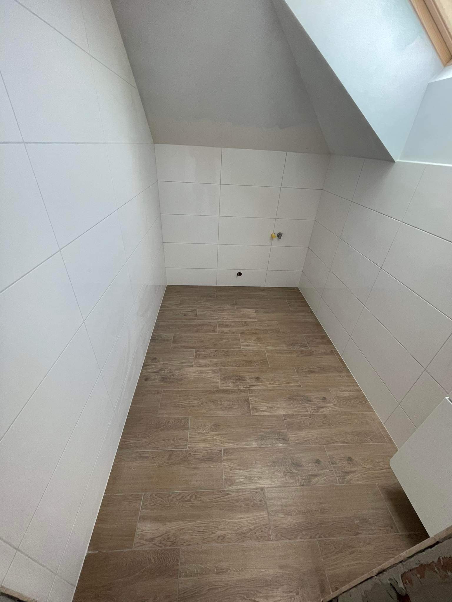 Wykończona łazienka na poddaszu z białymi płytkami ściennymi i podłogą imitującą drewno, widoczne przyłącza wodne.