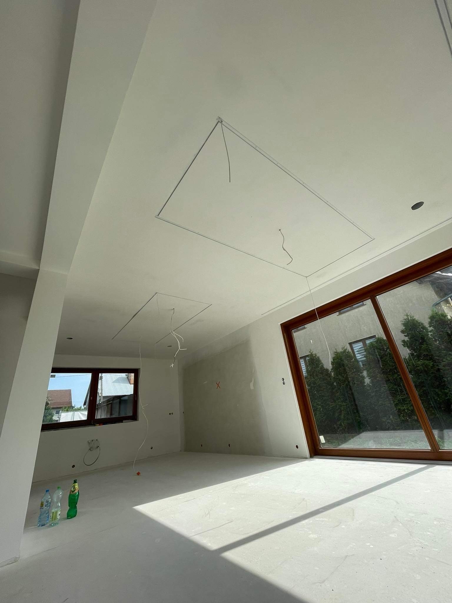 Surowe wnętrze pokoju z przygotowanymi otworami w suficie pod instalację, widoczne przewody, ściany po gładziowaniu, duże okno z widokiem na zieleń, butelki z wodą na podłodze.