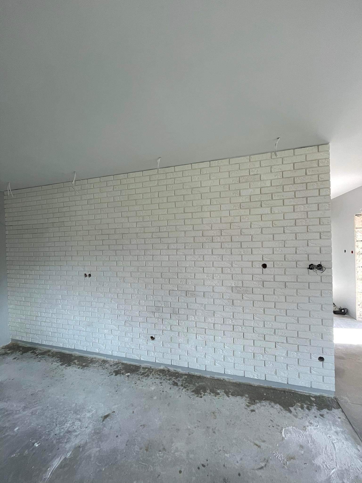 Świeżo położona, biała cegła dekoracyjna na ścianie działowej w surowym wnętrzu, widoczne otwory na gniazdka i wystające kable, surowy beton na podłodze.