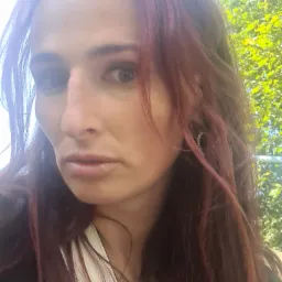 Portret kobiety z długimi, czerwonymi włosami, w tle zielone liście drzew, z kolczykiem w kształcie koła.