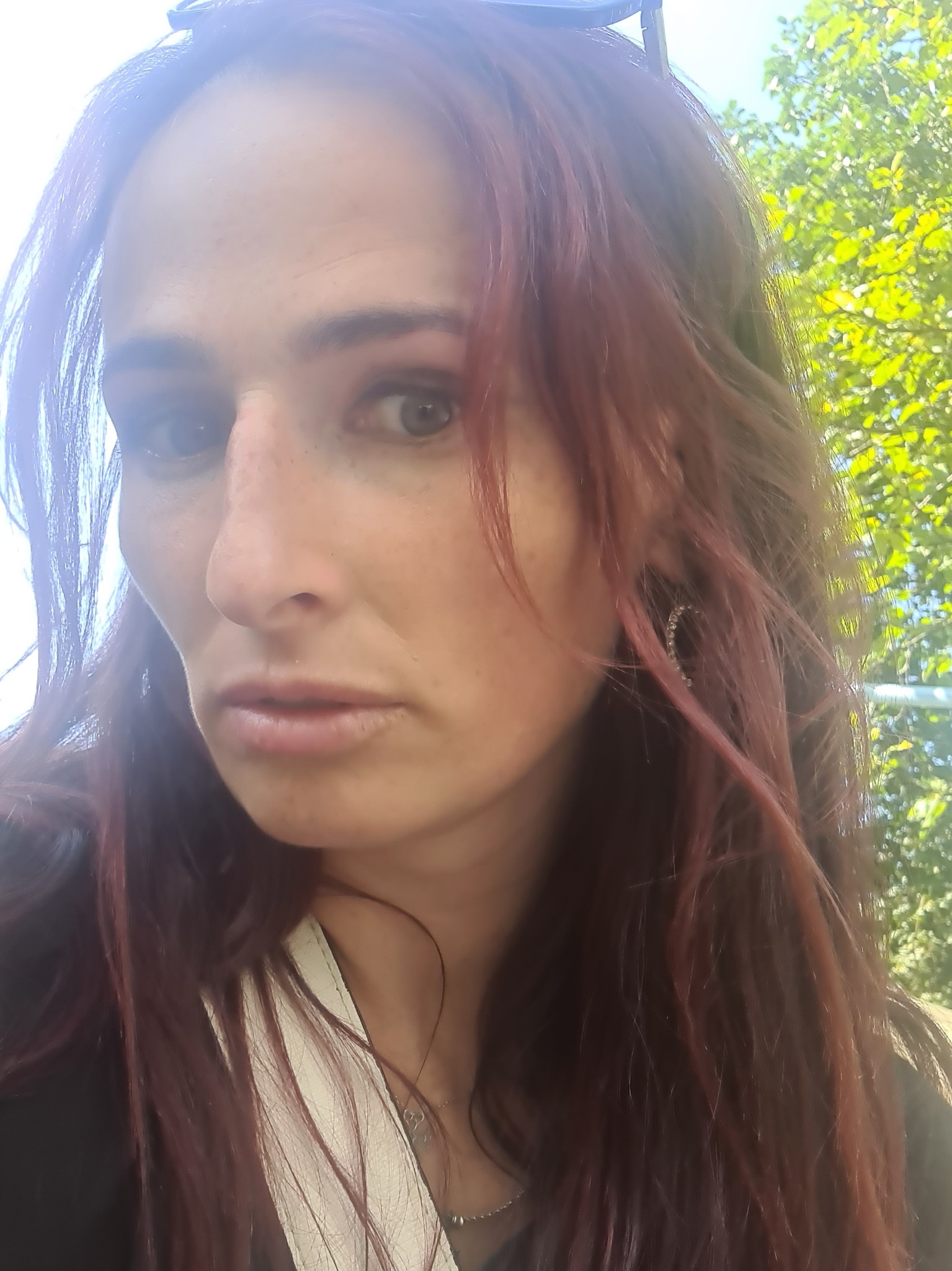 Portret kobiety z długimi, czerwonymi włosami, w tle zielone liście drzew, z kolczykiem w kształcie koła.