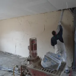Praca tynkarska w trakcie: mężczyzna w kombinezonie wyrównuje ścianę szpachelką. W tle agregat tynkarski i częściowo otynkowana ściana z instalacjami.