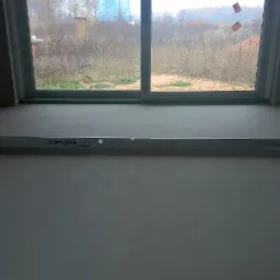 Wyrównana, szara wylewka w nowym pomieszczeniu z oknem na zewnątrz. Poziomica na posadzce. Widoczne gniazdka elektryczne i taśma malarska na ścianach.