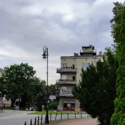 Ulica miejska z budynkami, drzewami i latarnią w pochmurny dzień. Na jednym z budynków widoczny maszt z polską flagą.