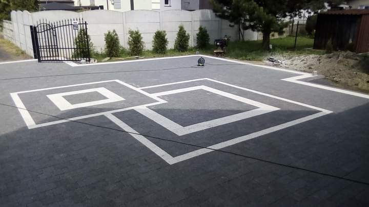 Ułożona, grafitowa kostka brukowa z białymi, geometrycznymi wzorami na podjeździe, widok z góry, brama w tle.