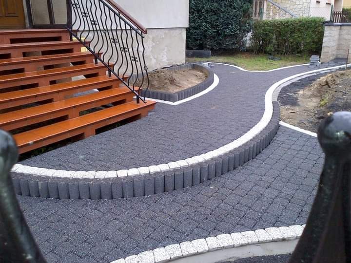 Ułożona, ciemnoszara kostka brukowa z białym obramowaniem tworząca ścieżkę i schody wejściowe do domu z drewnianą balustradą. Widoczny fragment metalowego ogrodzenia w dolnej części kadru.