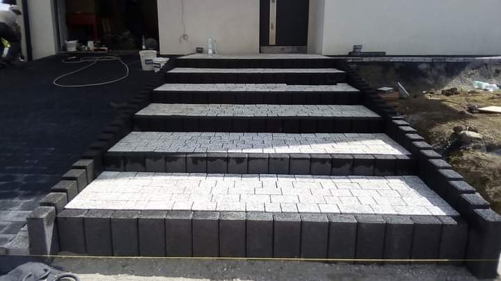 Schody wejściowe do domu wyłożone kostką brukową w odcieniach szarości i bieli, obramowane ciemnoszarą kostką, widoczne narzędzia i materiały budowlane w tle.