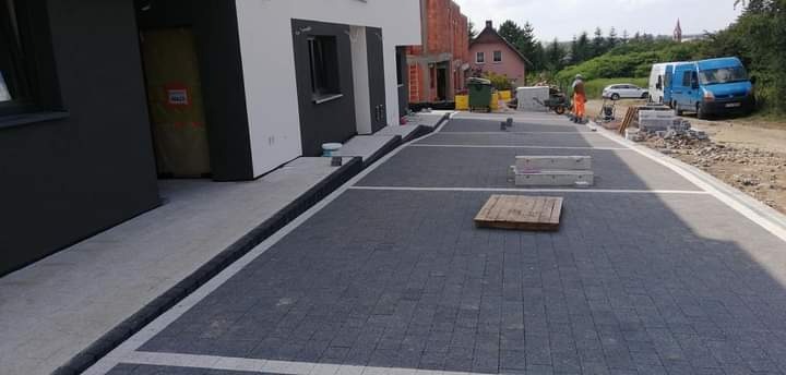 Ułożona kostka brukowa na podjeździe z wyznaczonymi miejscami parkingowymi, widoczny budynek w trakcie budowy i pracownik w oddali.