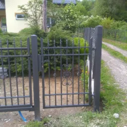 Otwarta, metalowa brama wjazdowa w kolorze grafitowym, z ozdobnymi elementami w kształcie serc, prowadząca na żwirową drogę, na tle domu z żółtą elewacją i zieleni.