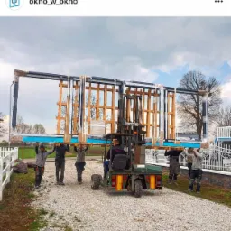 Pracownicy i wózek widłowy transportują pakiet okien w folii ochronnej na zewnątrz budynku, widoczne drewniane przekładki i metalowe ramy.