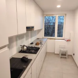 Nowoczesna, jasna kuchnia z białymi szafkami, szarym blatem, czarnym zlewem i płytą gazową z czajnikiem, widok na okno z drzewami, płytki podłogowe w odcieniach beżu, mały stołek.