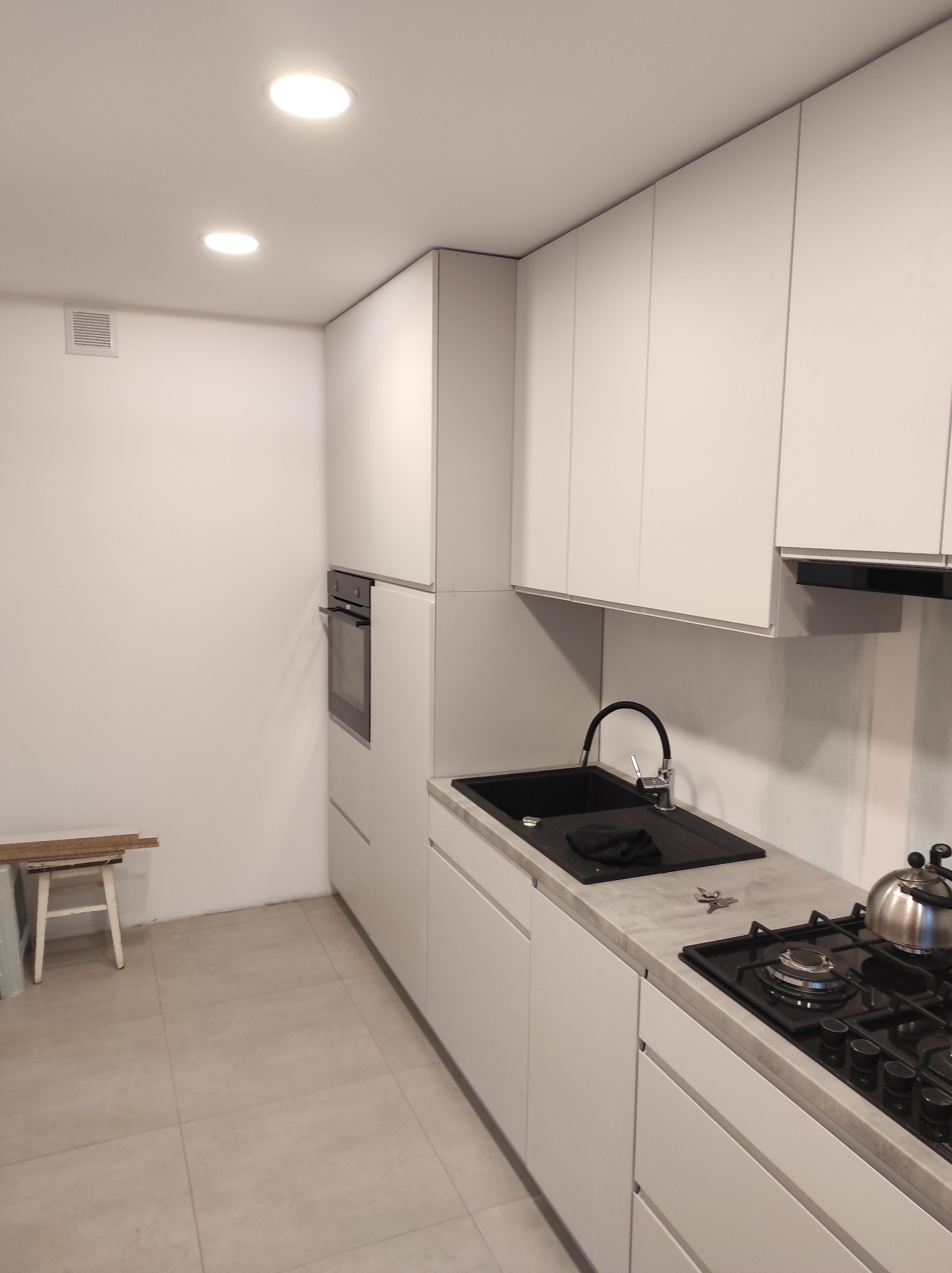 Nowoczesna, minimalistyczna kuchnia z białymi szafkami, czarnym zlewem i płytą gazową. Na blacie leżą klucze, a obok zlewu znajduje się czarna ścierka. W tle widoczne białe ściany i jasna podłoga...