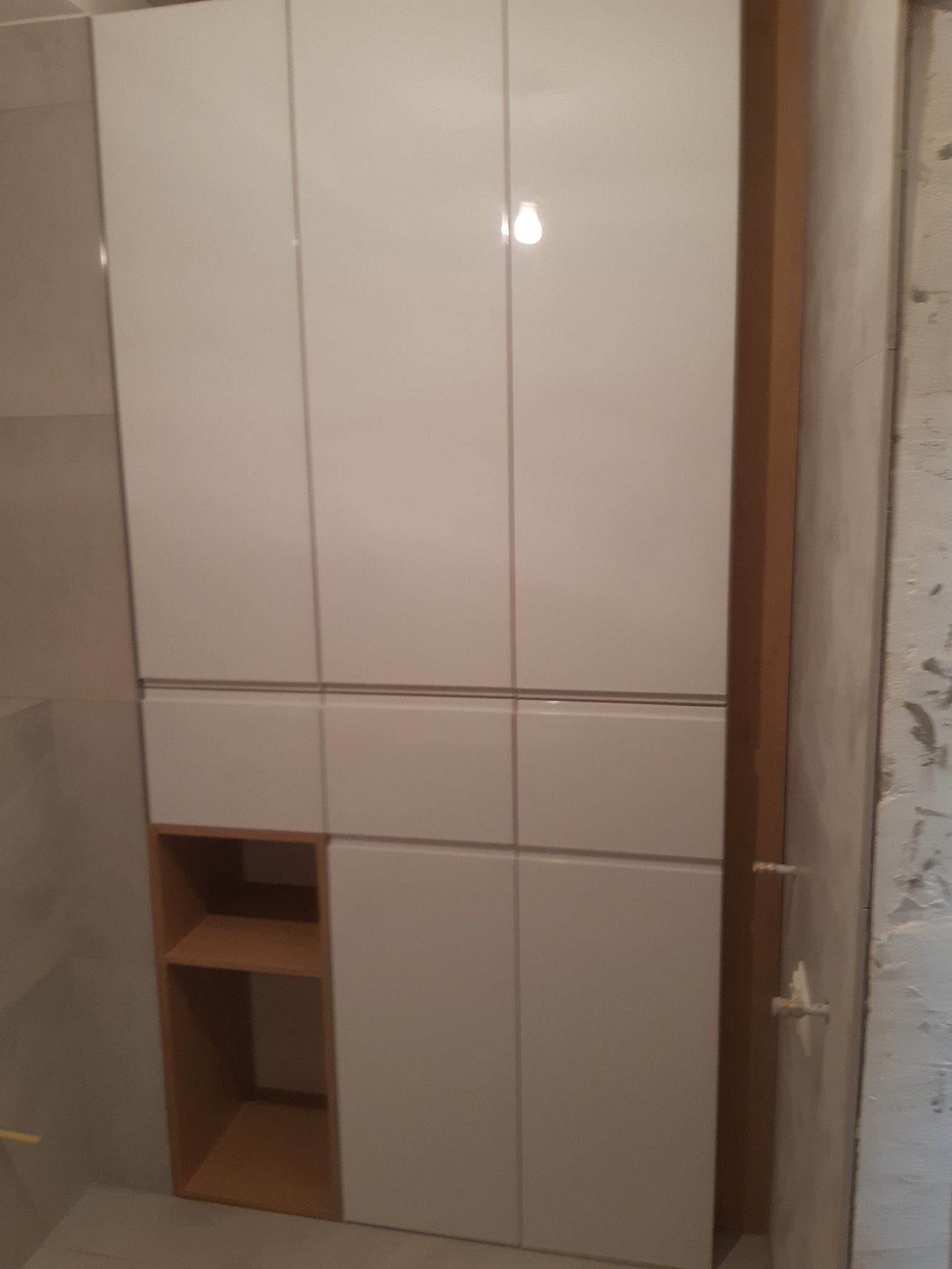 Biała szafka łazienkowa z połyskiem, z otwartą półką z jasnego drewna, zamontowana przy szarej ścianie wykończonej płytkami, widoczny fragment surowej ściany obok.