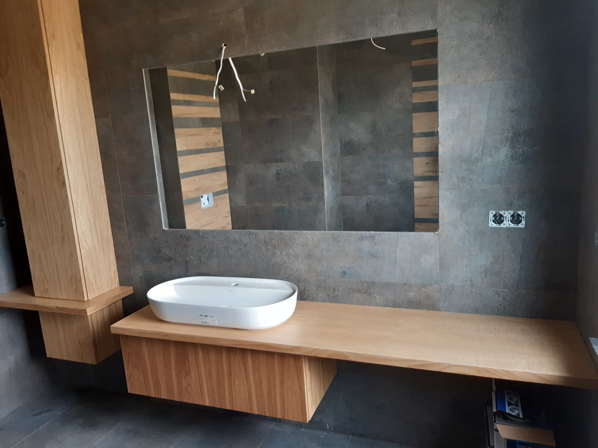 Nowoczesna łazienka z umywalką nablatową na drewnianym blacie, wisząca szafka z jasnego drewna, szare płytki na ścianach, widoczne przewody elektryczne i odbicie w lustrze
