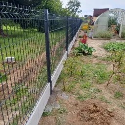 Nowoczesne, metalowe ogrodzenie panelowe w kolorze grafitowym, osadzone na betonowej podmurówce, oddzielające ogródek z warzywami i kwiatami od reszty posesji, widok na szklarnię w tle.