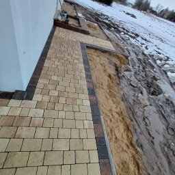 Prec-Bud - Fragment chodnika z kostki brukowej w odcieniach beżu i brązu, ułożonego przy białej ścianie budynku, z widocznym przygotowanym podłożem i resztkami śniegu w tle.