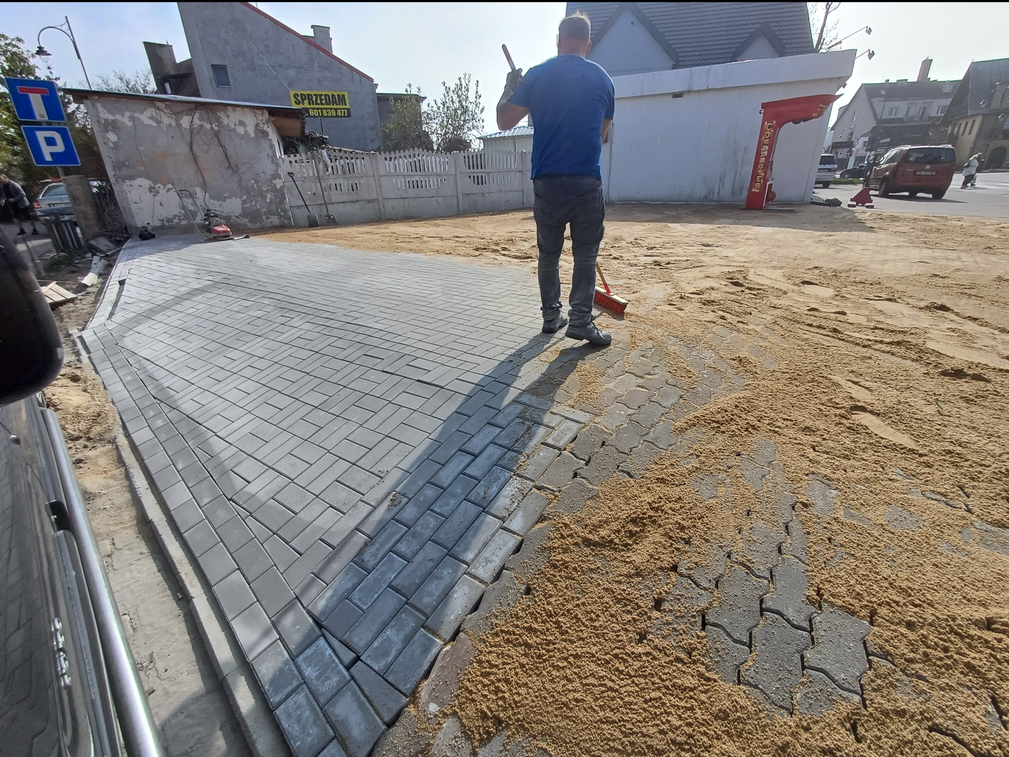 Układanie kostki brukowej: robotnik zamiata piasek z nowo ułożonej szarej kostki brukowej na podjeździe, widoczny fragment samochodu i budynki w tle.
