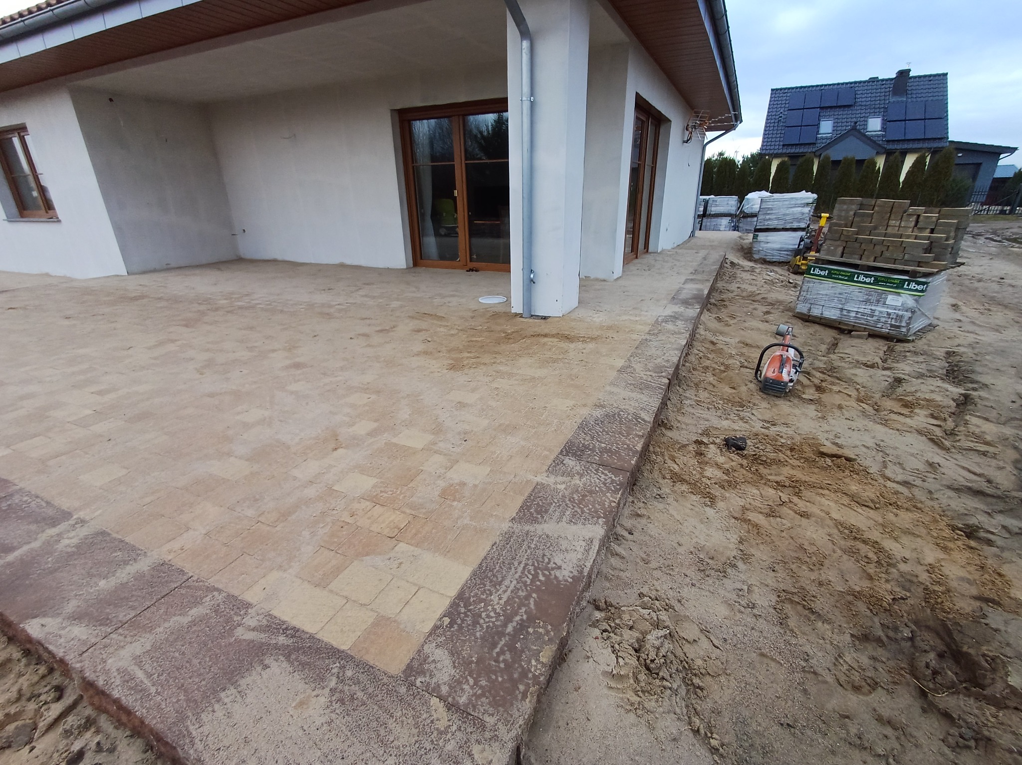 Wykończony taras z kostki brukowej w Kołobrzegu, z obrzeżem w ciemniejszym odcieniu, widoczne zapasy kostki i narzędzia na placu budowy, dom z panelami słonecznymi w tle.