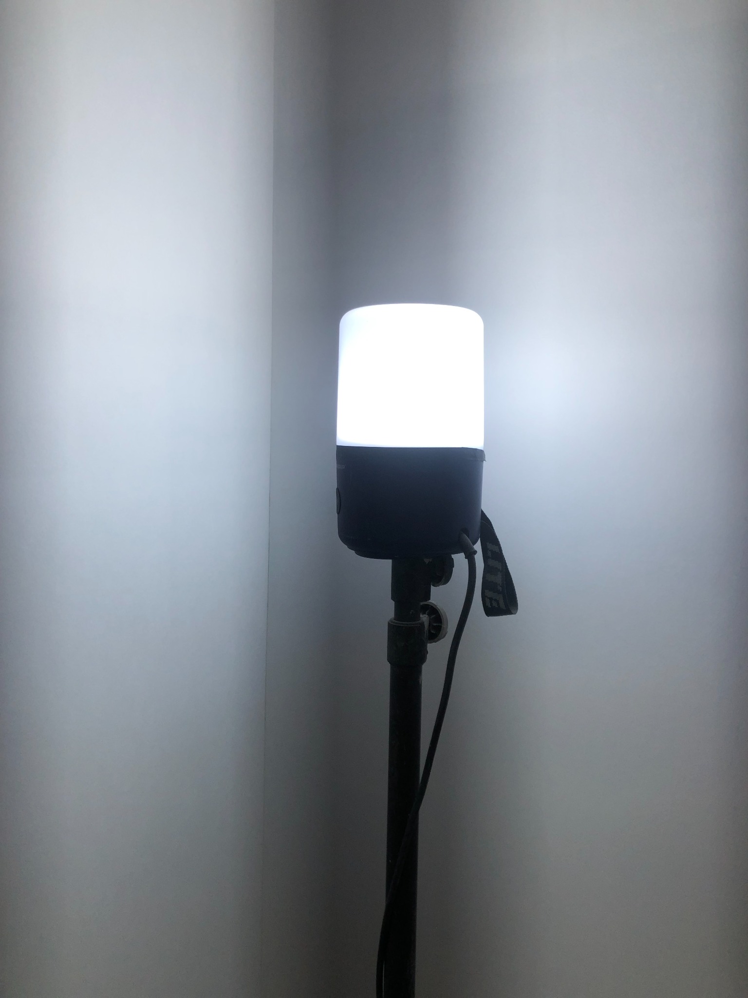 Lampa budowlana na statywie oświetlająca róg pomieszczenia z pomalowanymi ścianami, widoczne przewody i elementy regulacyjne statywu.