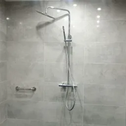 Wykończona kabina prysznicowa z szarymi płytkami, deszczownicą, uchwytem dla niepełnosprawnych i półką z kosmetykami.