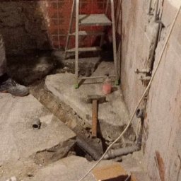 GRZEGORZ NOGA FIRMA HETMAN - Demontaż starej posadzki w łazience: widoczne fragmenty płytek, gruz, rura kanalizacyjna, drabina aluminiowa oraz narzędzia - młotek i szpachelka.