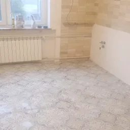 Wykończona podłoga w kuchni z ułożonymi płytkami w odcieniach szarości, częściowo wykafelkowana ściana, grzejnik pod oknem, przygotowane materiały budowlane na parapecie.