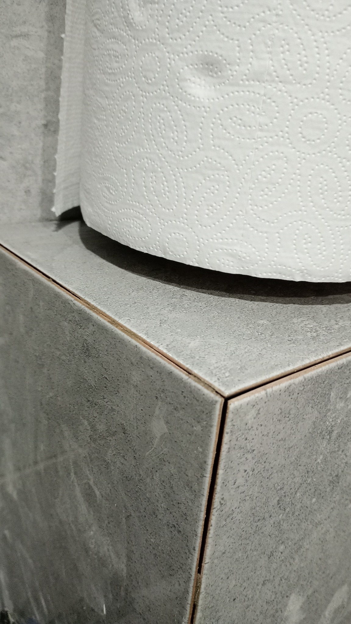 Szare, matowe płytki ceramiczne ułożone na narożniku, z widocznymi fugami i rolką białego papieru toaletowego na górze.