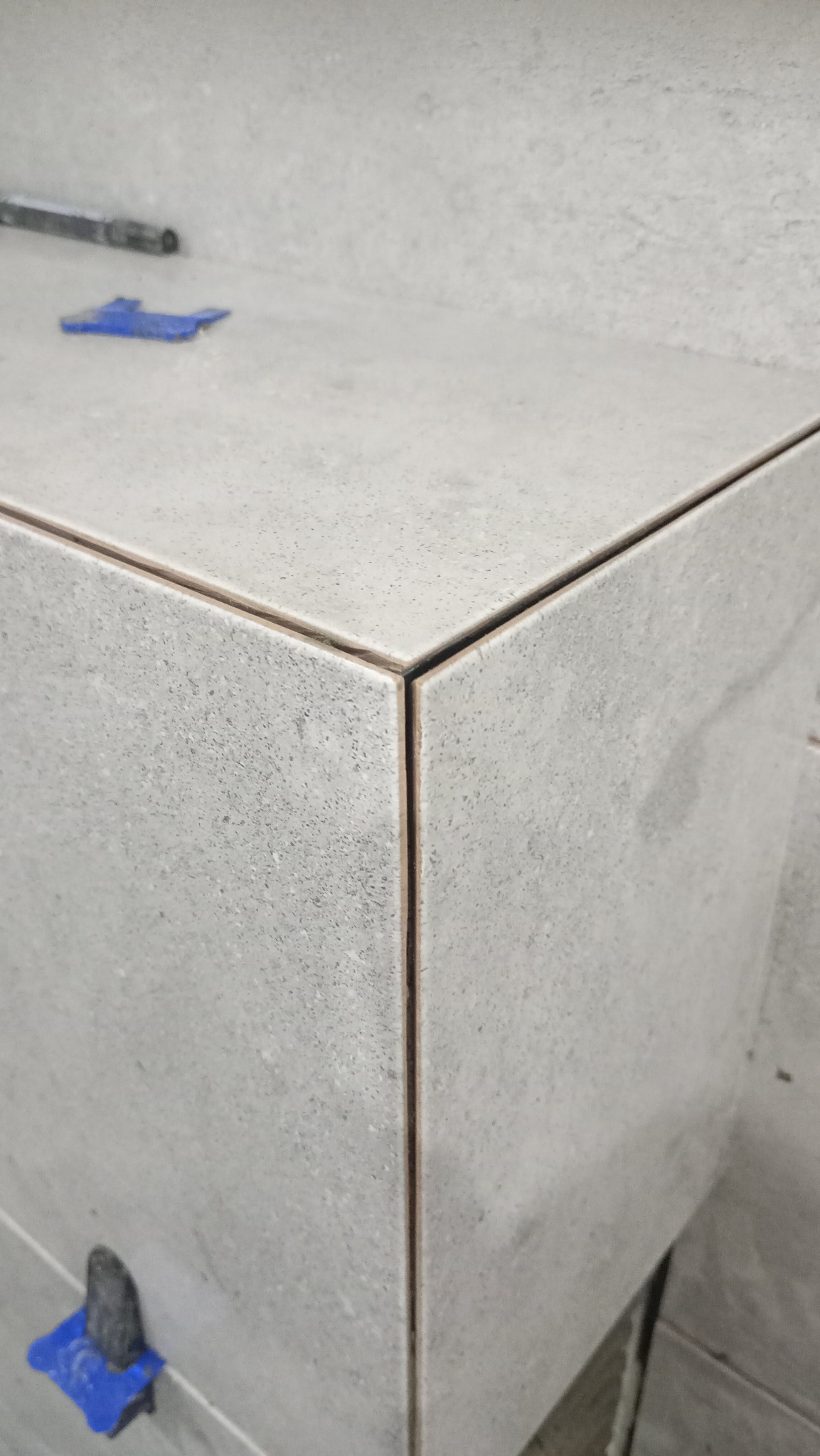 Szczegółowe ujęcie narożnika wykończonego szarymi płytkami z widocznymi plastikowymi klinami poziomującymi i fugą w kolorze brązowym.