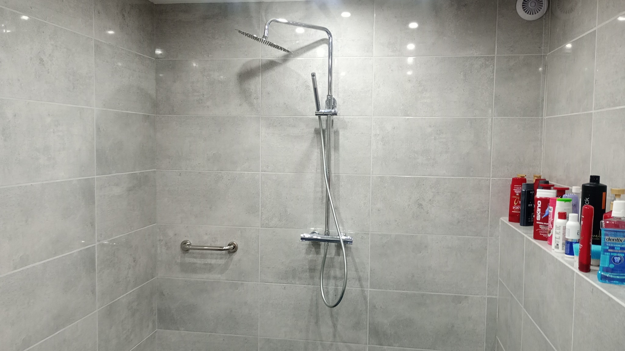 Wykończona kabina prysznicowa z szarymi płytkami, deszczownicą, uchwytem dla niepełnosprawnych i półką z kosmetykami.