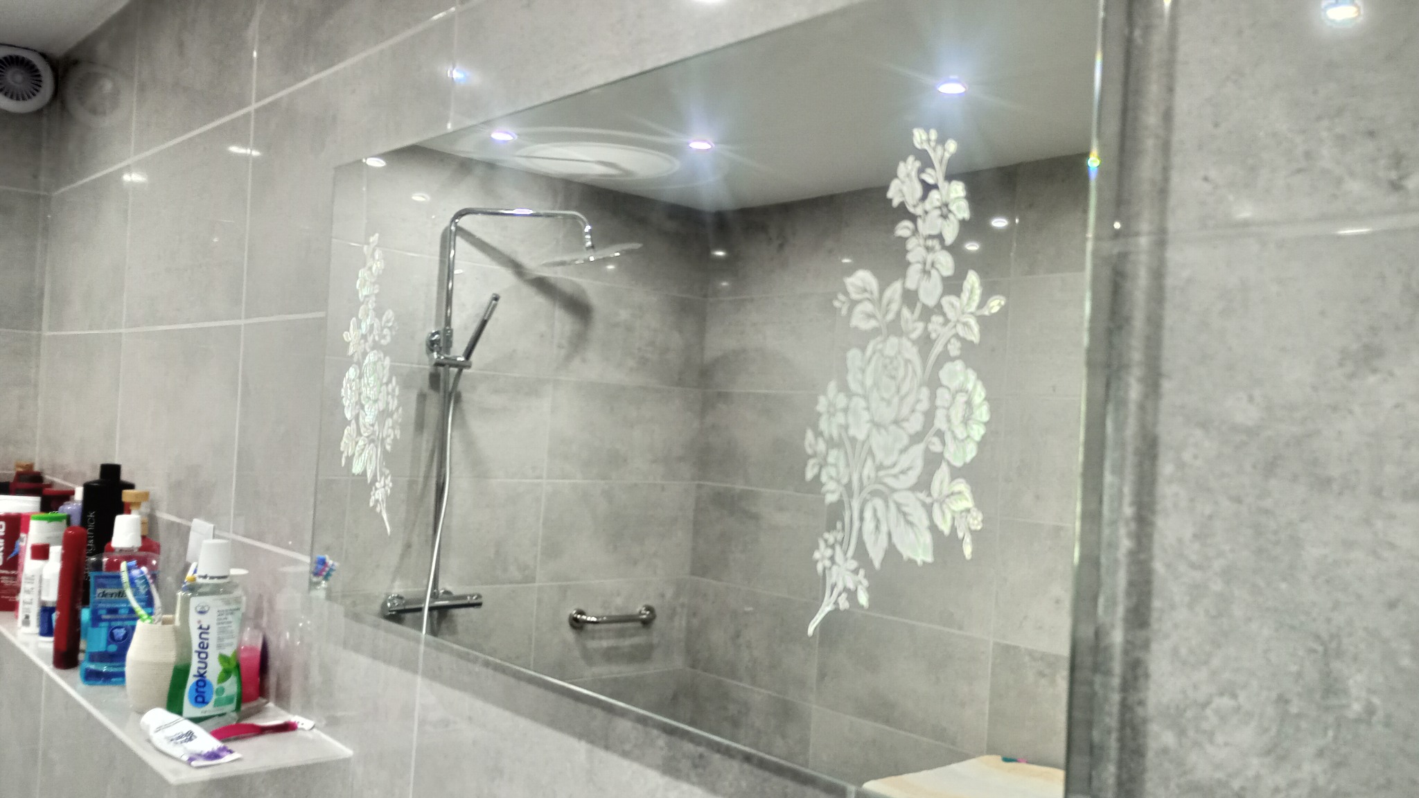 Szara łazienka z prysznicem, lustrem z motywem kwiatowym i półką z kosmetykami, widoczna wentylacja sufitowa.