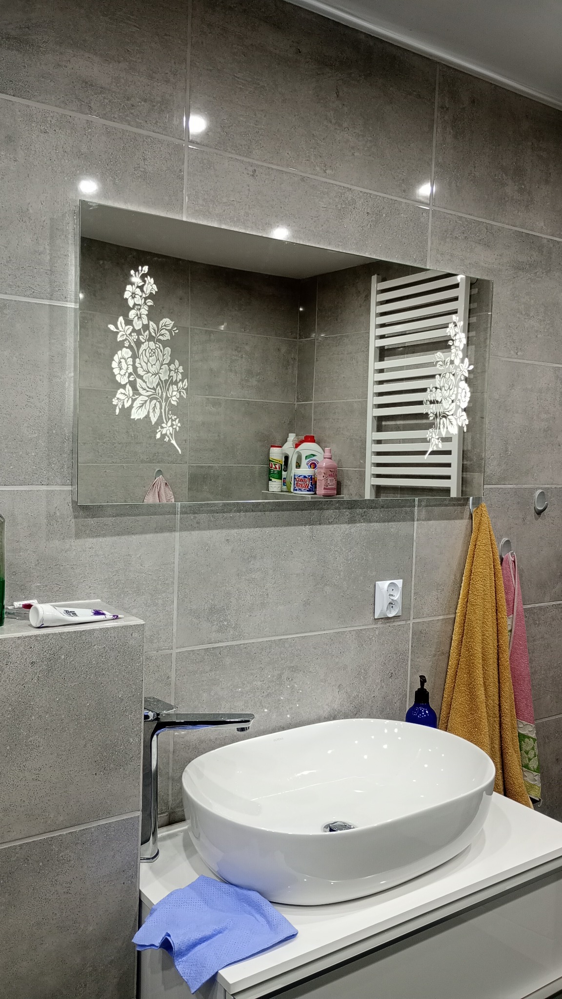 Szara łazienka z owalną umywalką nablatową, chromowaną baterią i lustrem z motywem kwiatowym, odbijającym grzejnik drabinkowy i półkę z kosmetykami.