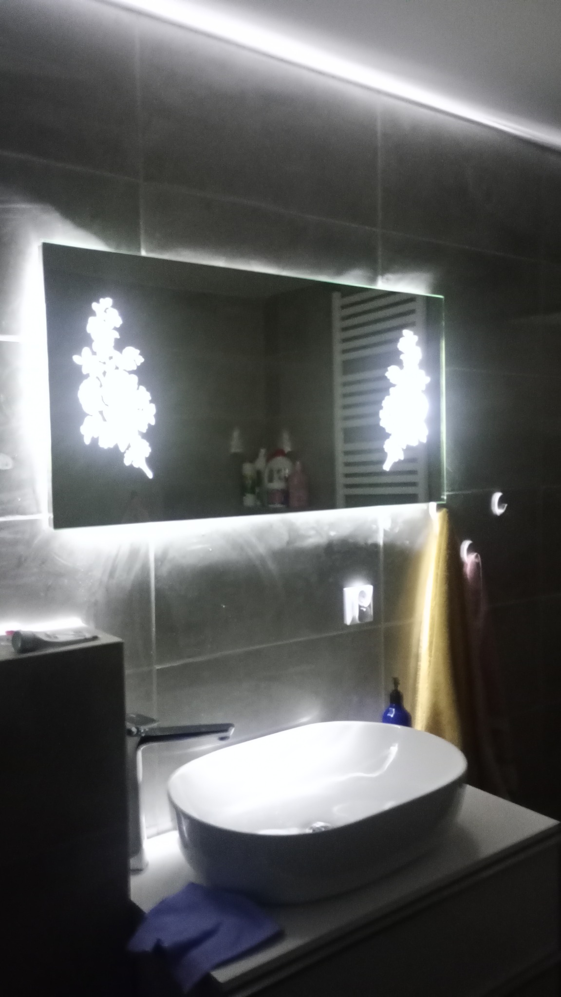 Nowoczesna łazienka z umywalką nablatową, podświetlanym lustrem z motywem kwiatowym i szarymi płytkami na ścianach.