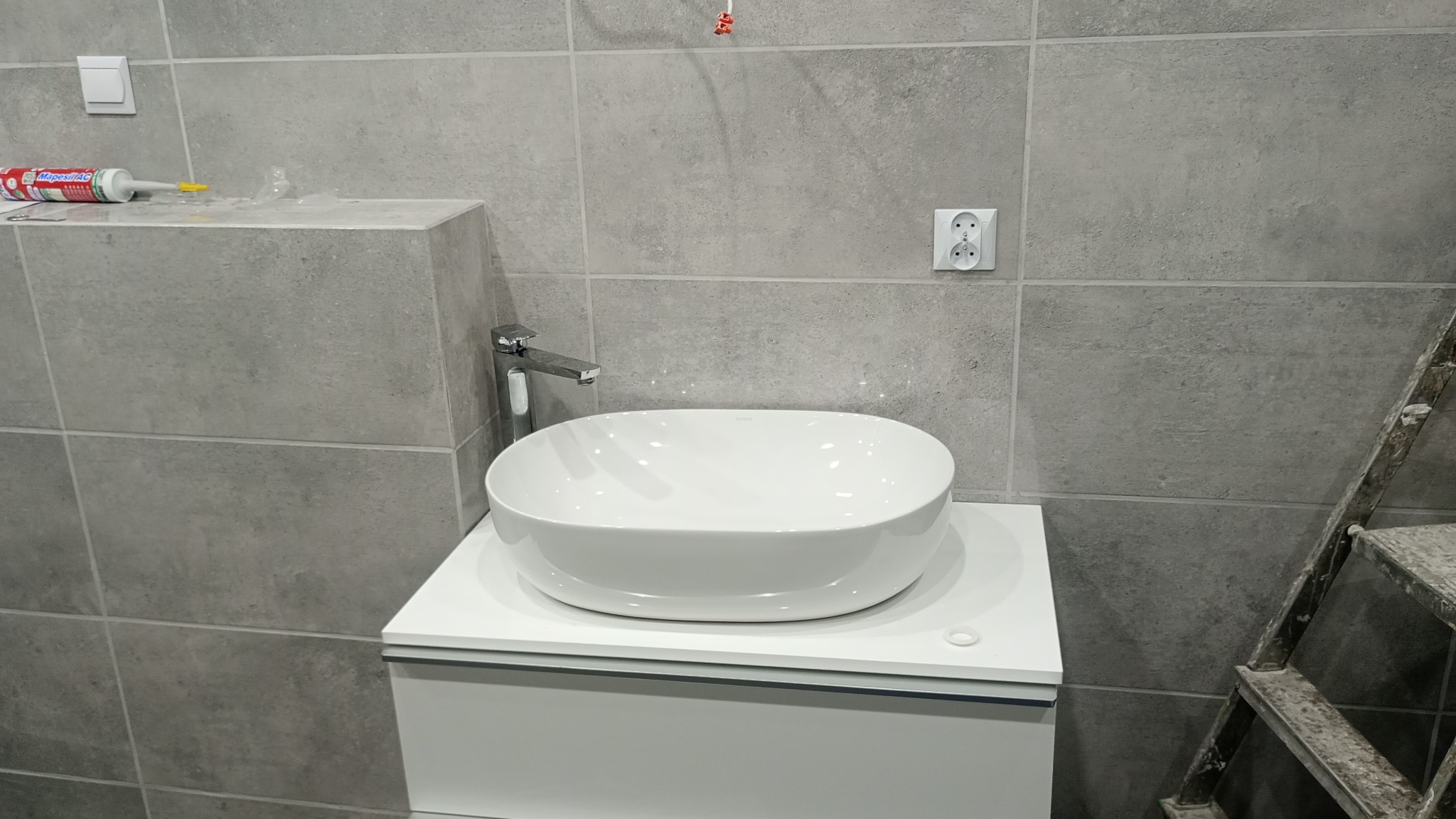 Montaż umywalki nablatowej na szafce w łazience wykończonej szarymi płytkami, widoczna drabina malarska, silikon i gniazdko elektryczne.