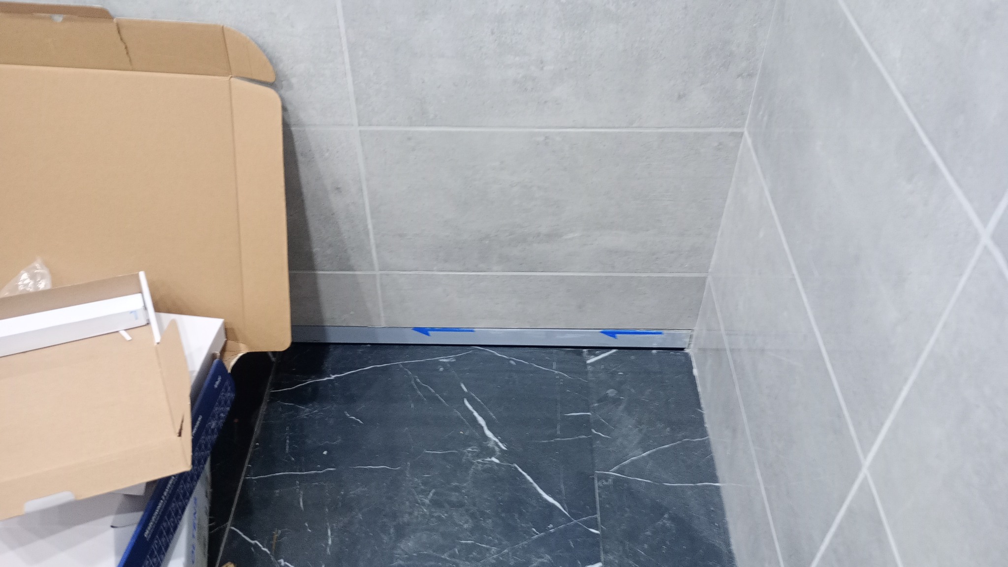 Fragment łazienki w trakcie wykańczania: szare płytki ścienne, czarna marmurowa podłoga, listwa przypodłogowa z niebieską folią ochronną, kartonowe pudełka z materiałami budowlanymi w rogu.
