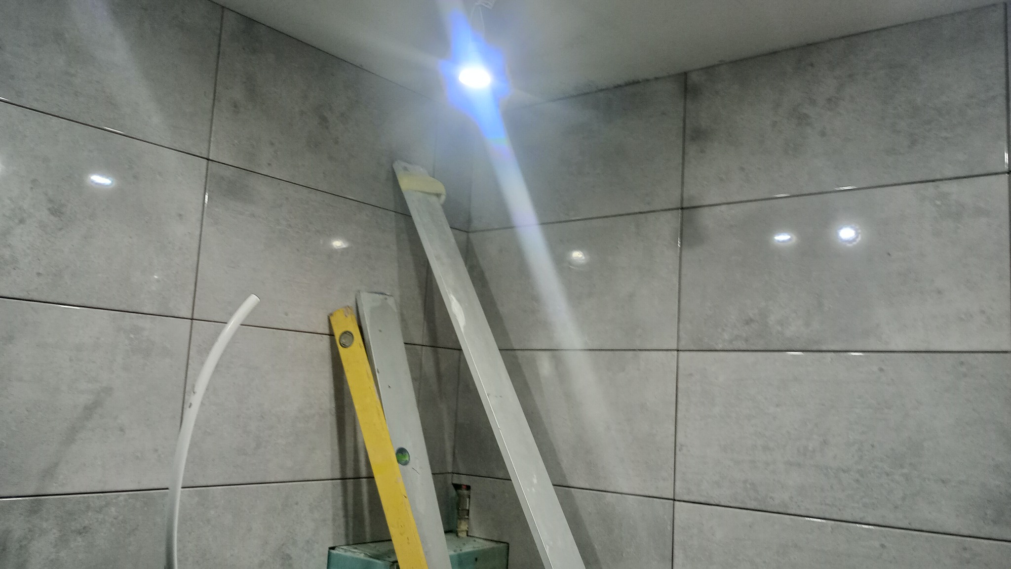 Świeżo ułożone szare płytki w łazience, widoczne poziomice i rurka instalacyjna przy ścianie, intensywne światło górne.