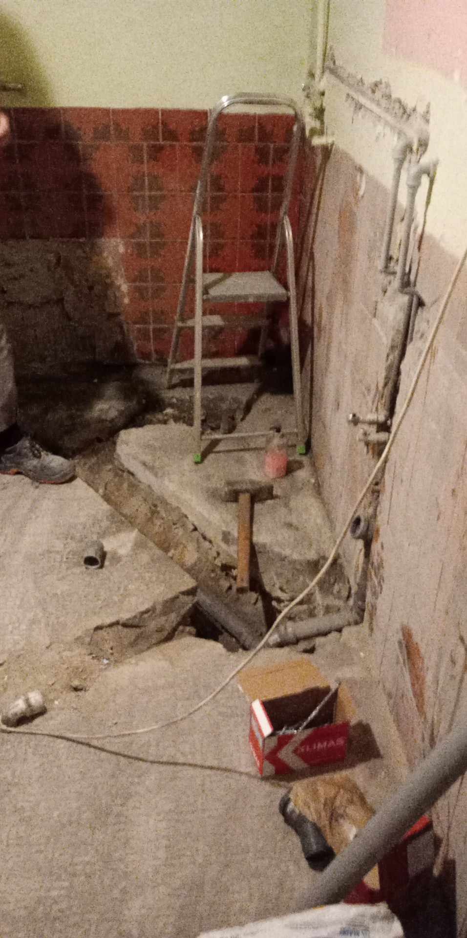 Demontaż starej posadzki w łazience: widoczne fragmenty płytek, gruz, rura kanalizacyjna, drabina aluminiowa oraz narzędzia - młotek i szpachelka.