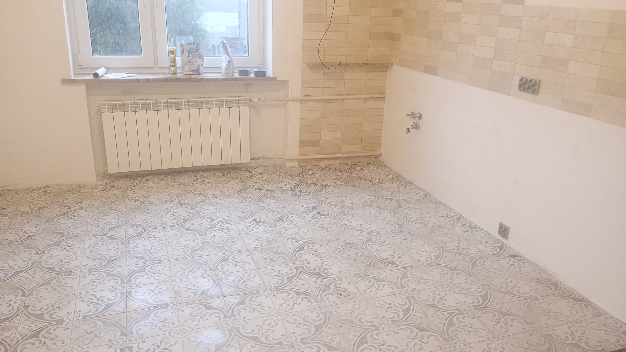Wykończona podłoga w kuchni z ułożonymi płytkami w odcieniach szarości, częściowo wykafelkowana ściana, grzejnik pod oknem, przygotowane materiały budowlane na parapecie.