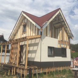 Indywidualny przedsiebiorca - Budowa domu w technologii szkieletowej: widoczna konstrukcja drewniana, częściowo ocieplona, z fragmentami elewacji i dachem z czerwonej dachówki, na pierwszym planie fragment tarasu w trakcie...