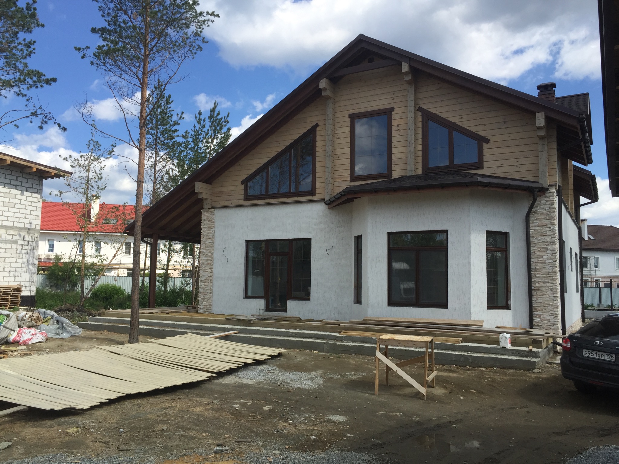 Dwukondygnacyjny dom w trakcie budowy z elementami drewnianymi i białym tynkiem, widoczne przygotowane deski i podłoże pod taras.