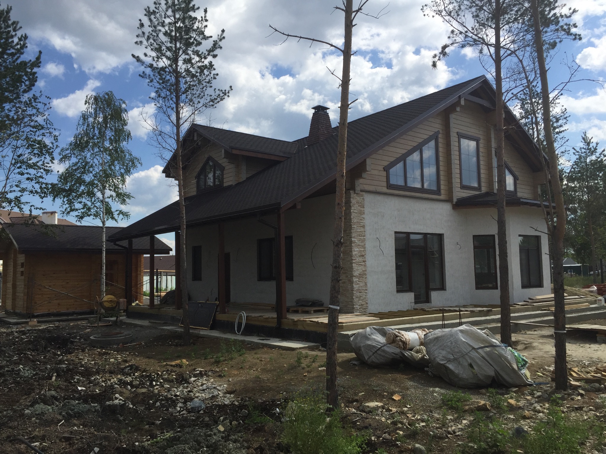 Dwukondygnacyjny dom w trakcie budowy z elementami drewnianymi i jasną elewacją, widoczne worki z materiałami budowlanymi i drzewa na pierwszym planie.